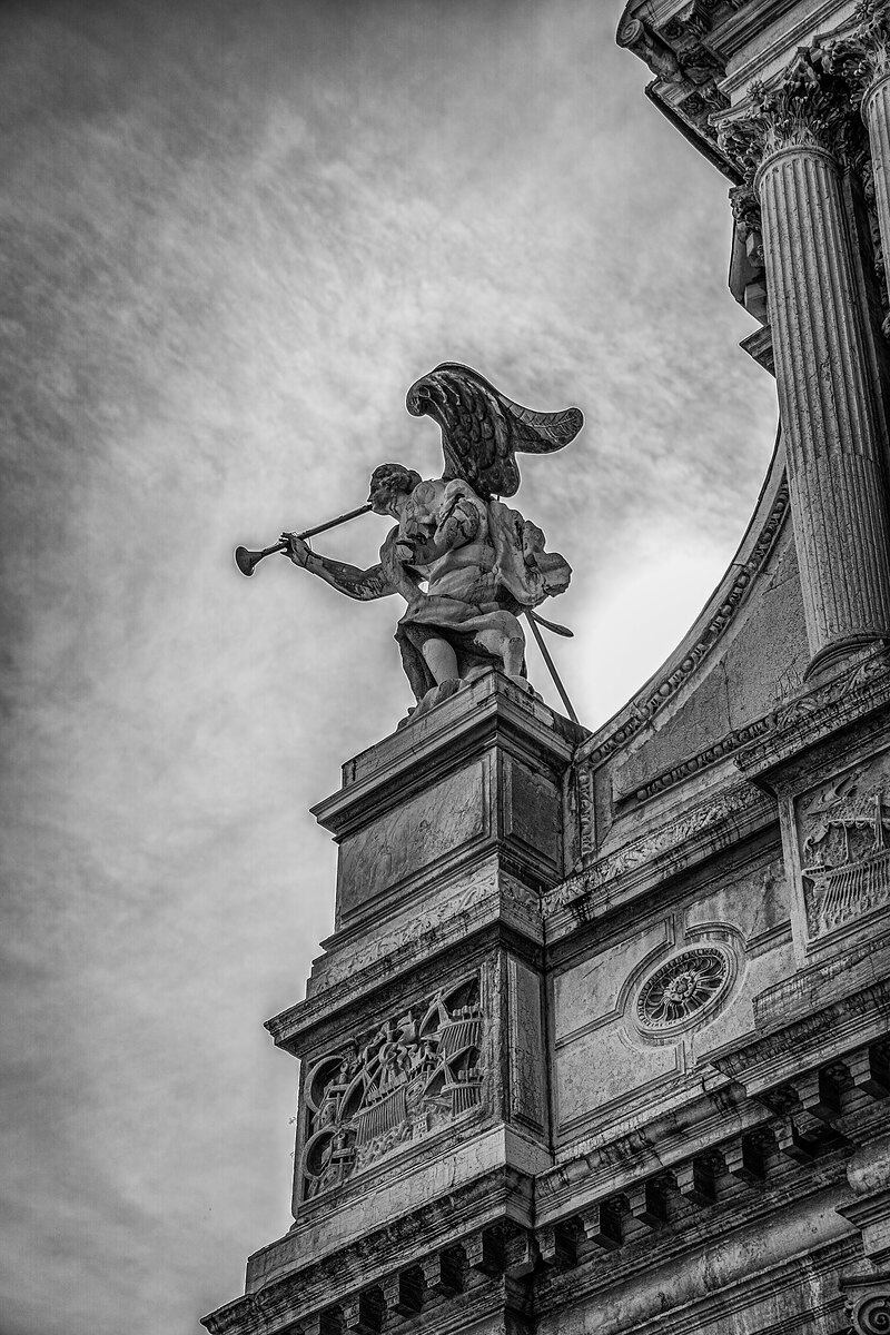 Black-and-white view of a trumpeting angel statue on an ornate church facade against a cloudy sky.