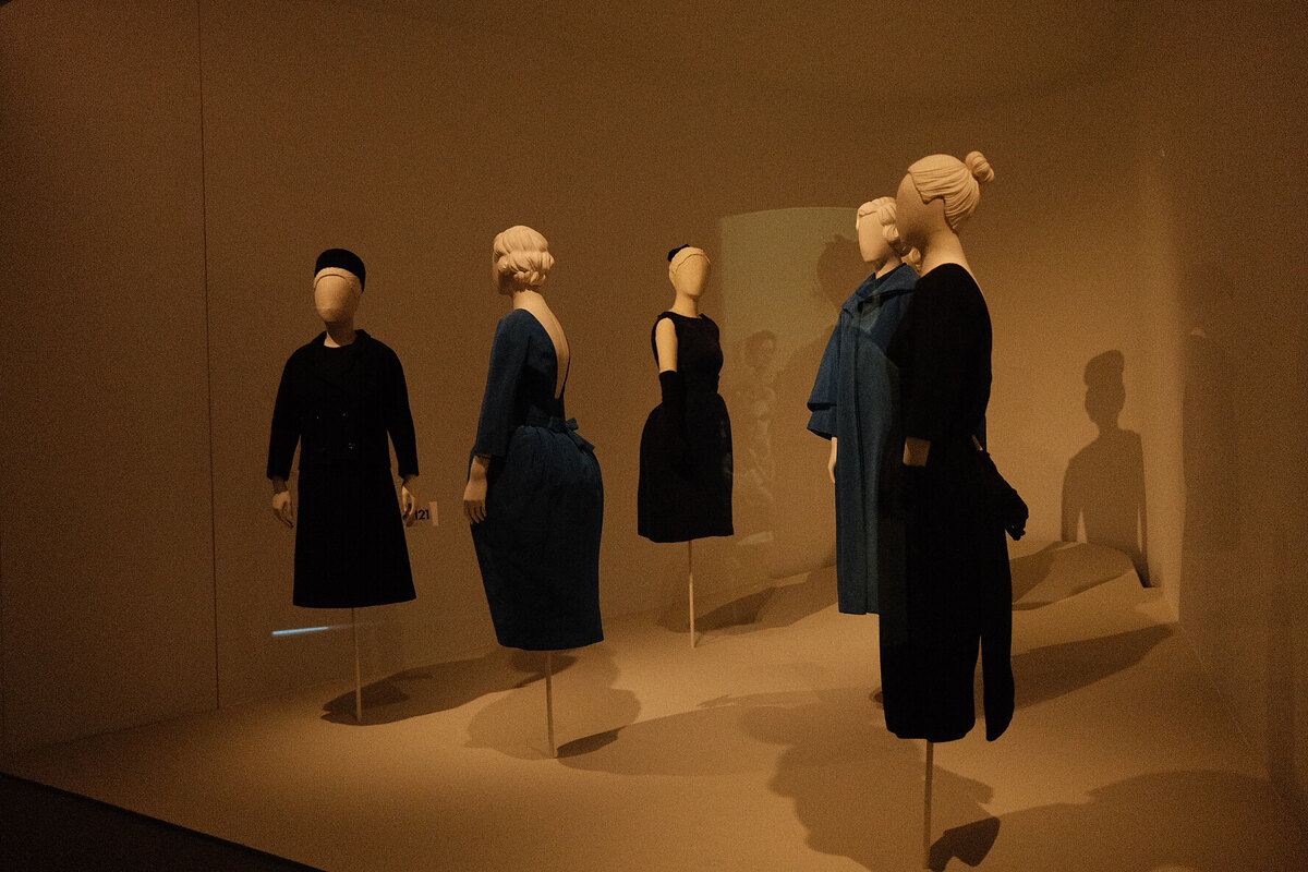 Five faceless white mannequins in black and blue dresses in a dim museum display case.