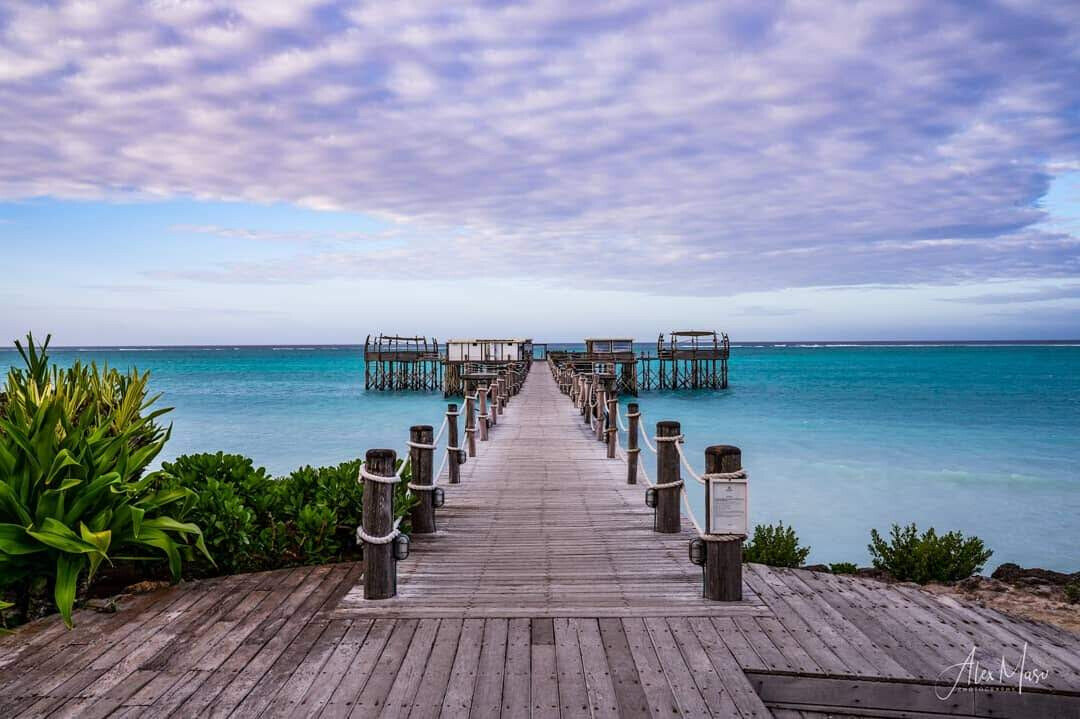 A wooden jetty on posts running out over turquoise water towards thatched overwater pavilions, under a sky of small high clouds.