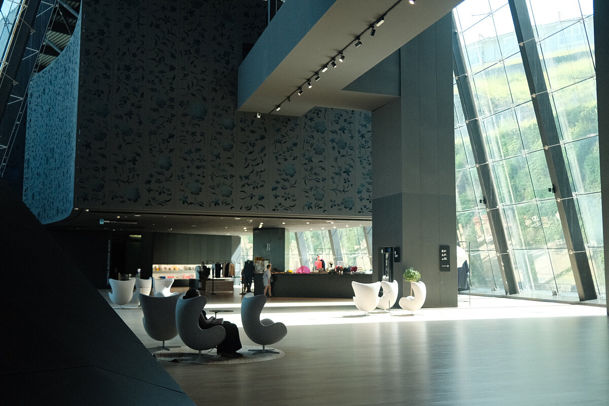 Spacious modern museum lobby with wood floor, glass windows, egg-shaped chairs and people at a counter.