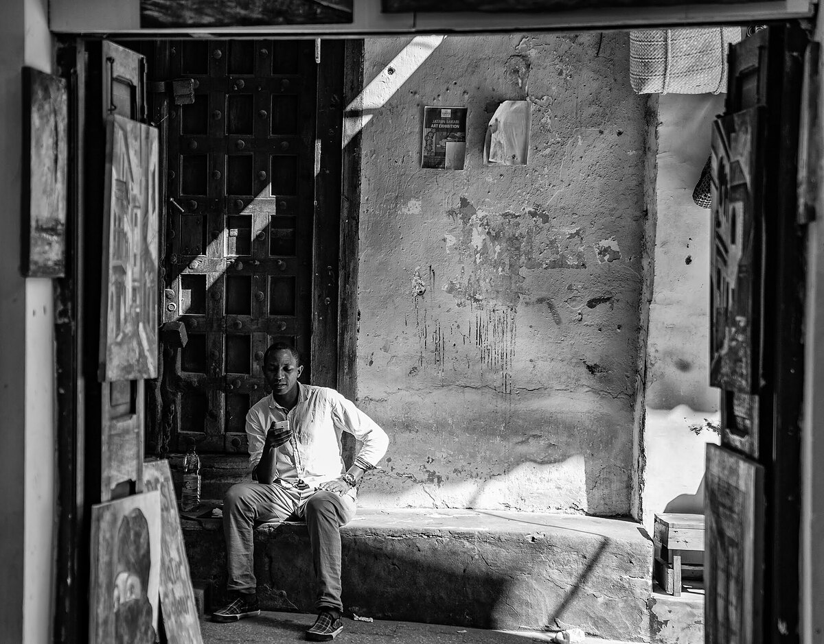 Black-and-white photograph of a man sitting on a shop step looking at his mobile phone, paintings leaning against the wall beside him, an old studded door behind him and hard sunlight across weathered plaster.