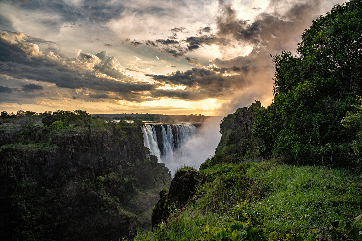 Wide view of Victoria Falls at sunrise - a wall of falling water and rising mist framed by green vegetation under a golden sky.