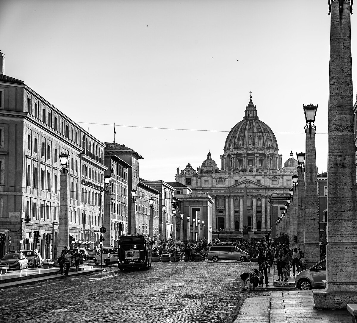 Black-and-white view down a wide Rome avenue toward the dome of St Peter's Basilica, with a bus, cars and crowds.