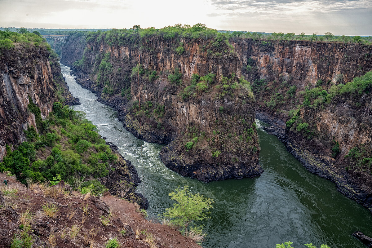 Looking down into a deep river gorge - the Zambezi River winding between dark cliffs toward a steel arch bridge in the distance.