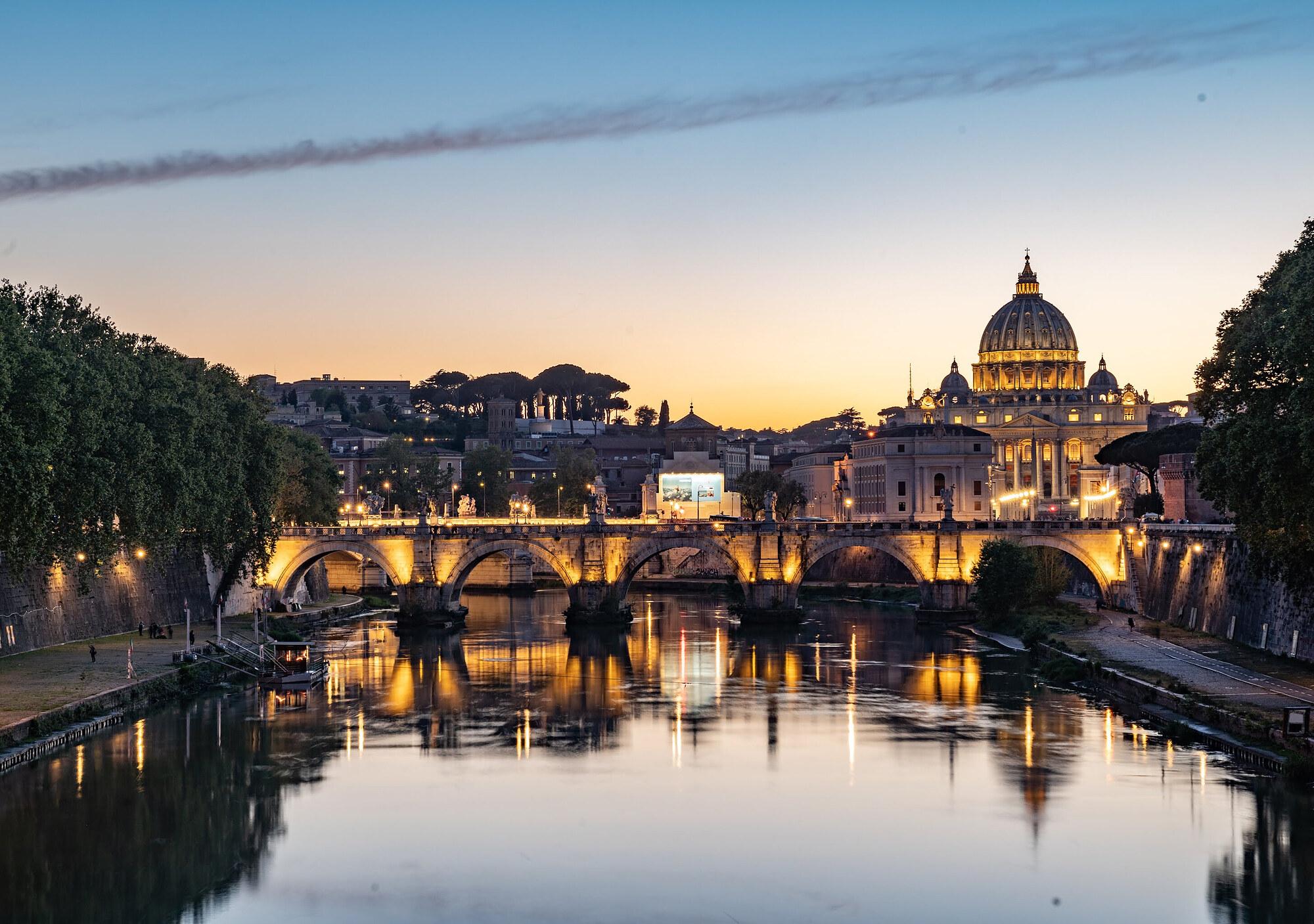 Dusk view across the River Tiber of an illuminated bridge with the floodlit dome of St Peter's Basilica beyond.