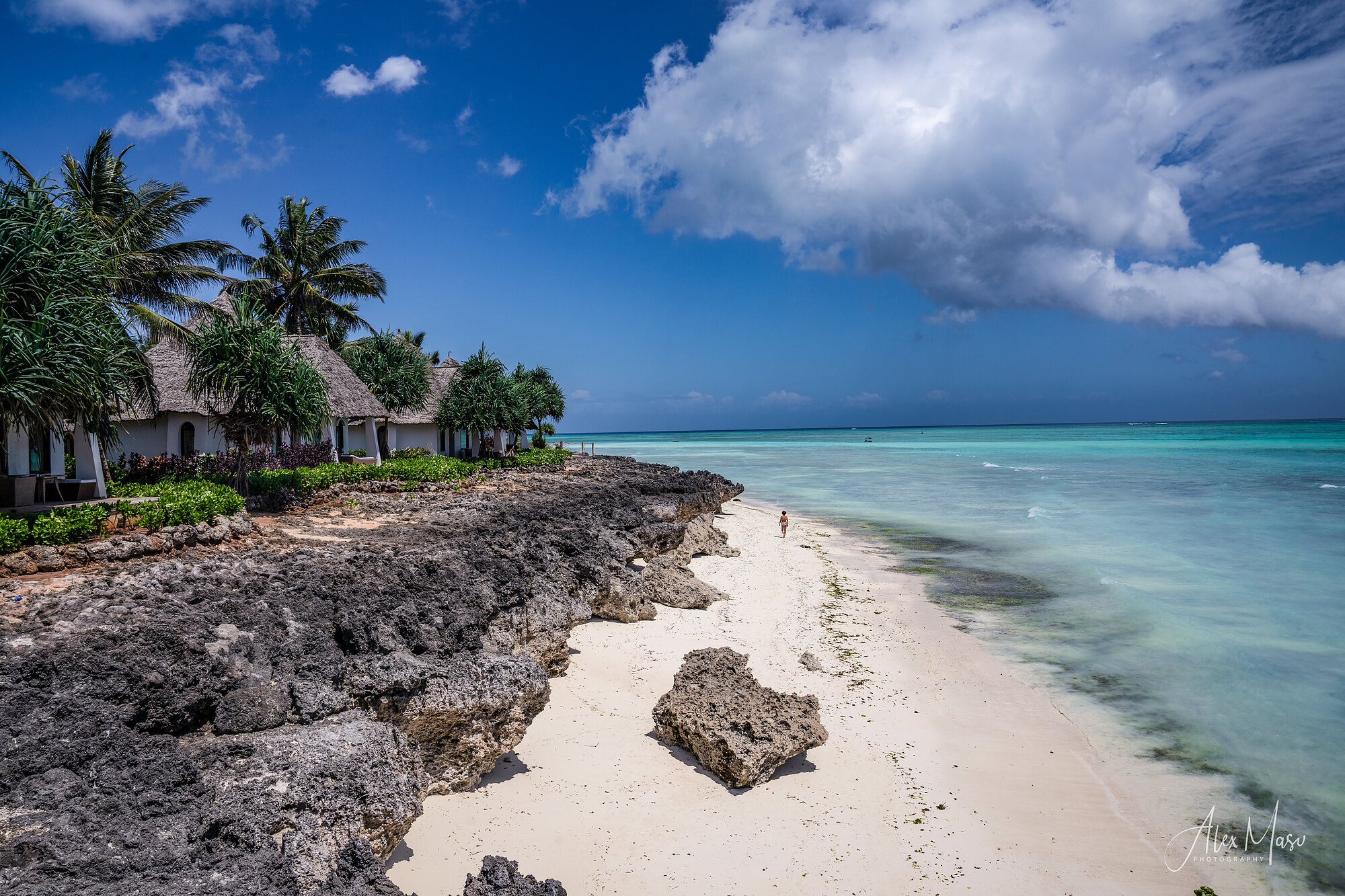 White thatched bungalows and palm trees on a coral cliff above a pale sand beach, with turquoise sea and a large cumulus sky, and one person walking in the shallows.