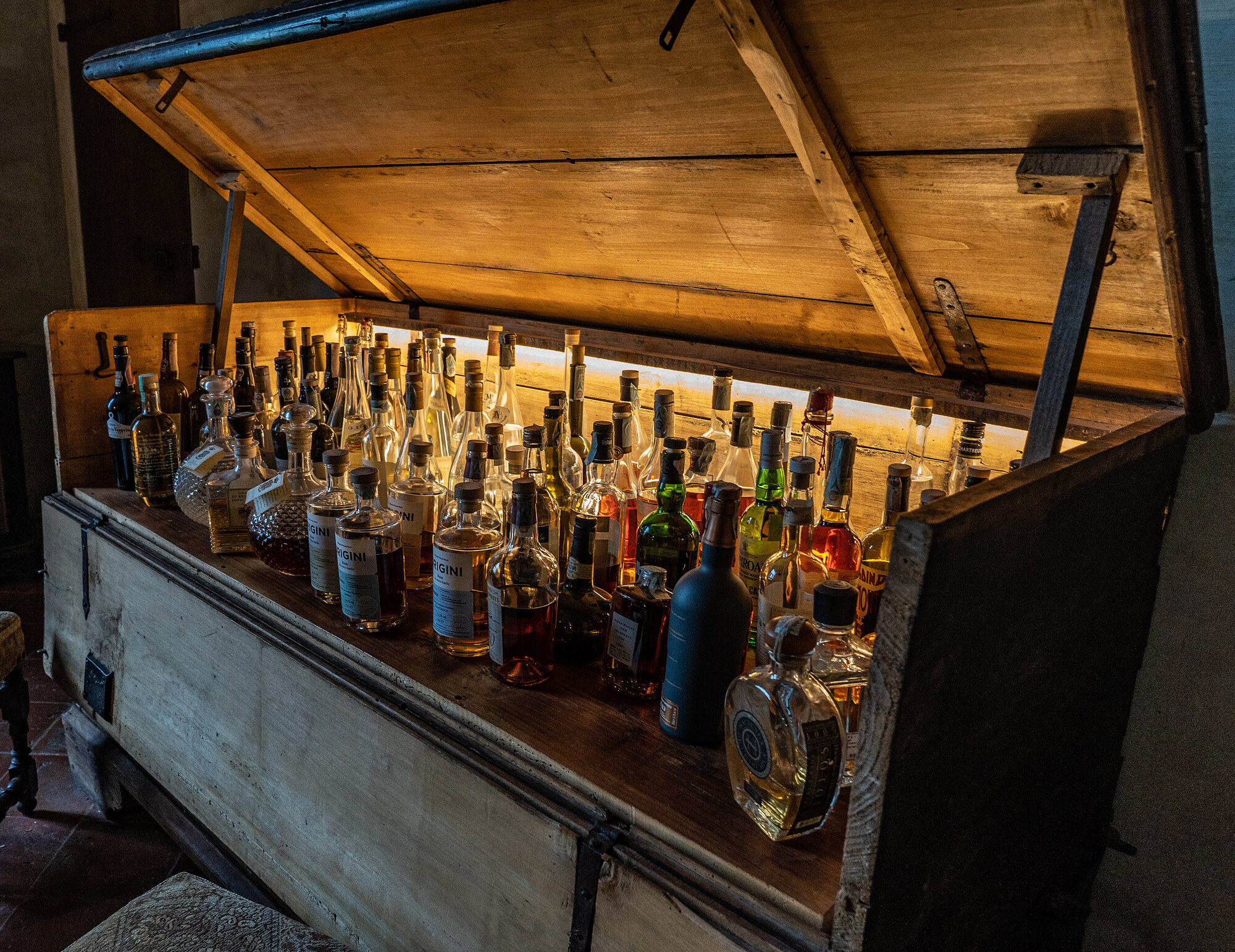 An open wooden chest filled with rows of glass spirit bottles lit warmly from behind.