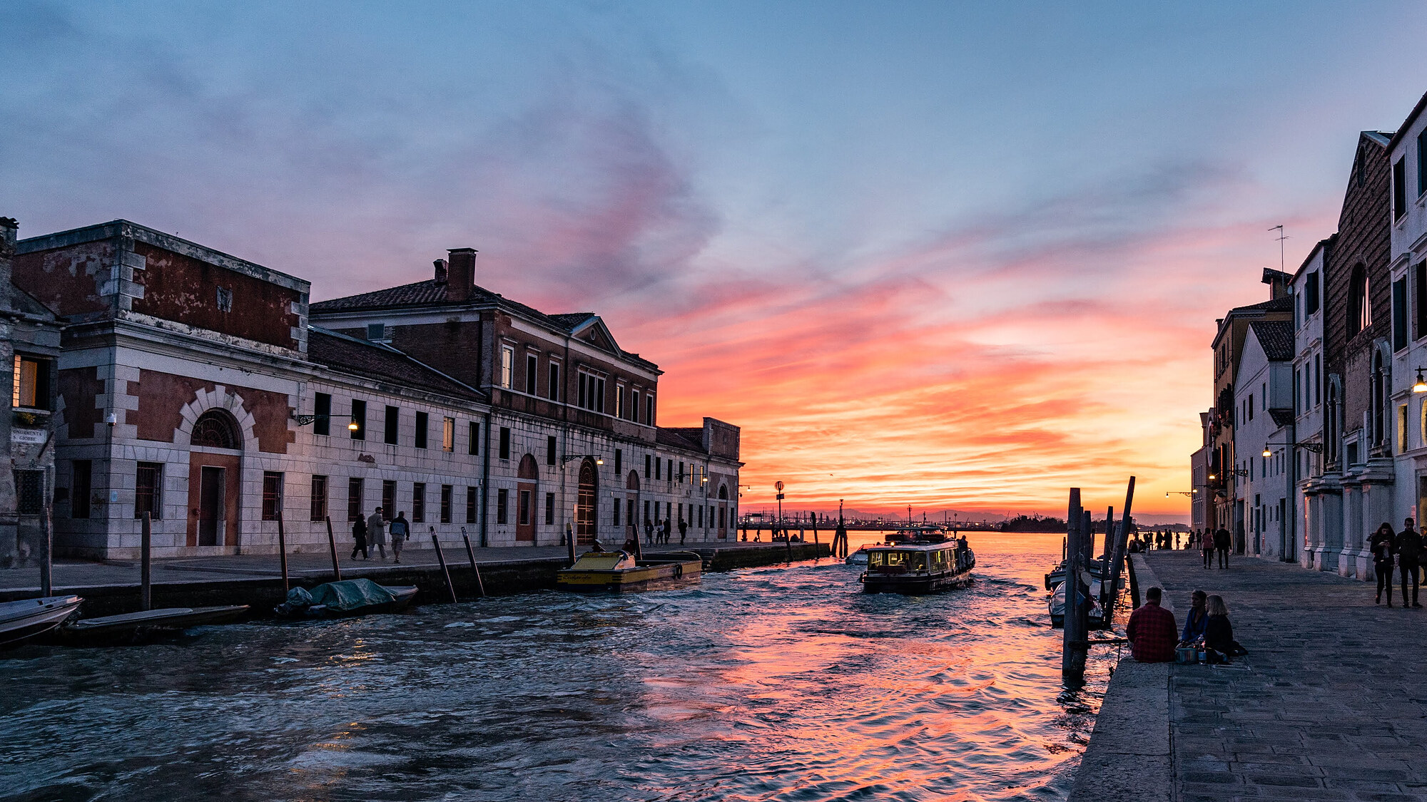 Sunset over a Venice canal with a passenger boat and historic buildings on both banks.