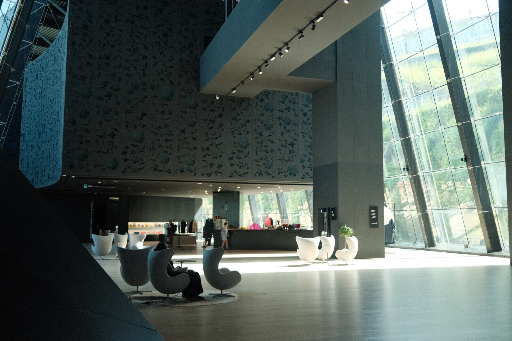 Spacious modern museum lobby with wood floor, glass windows, egg-shaped chairs and people at a counter.