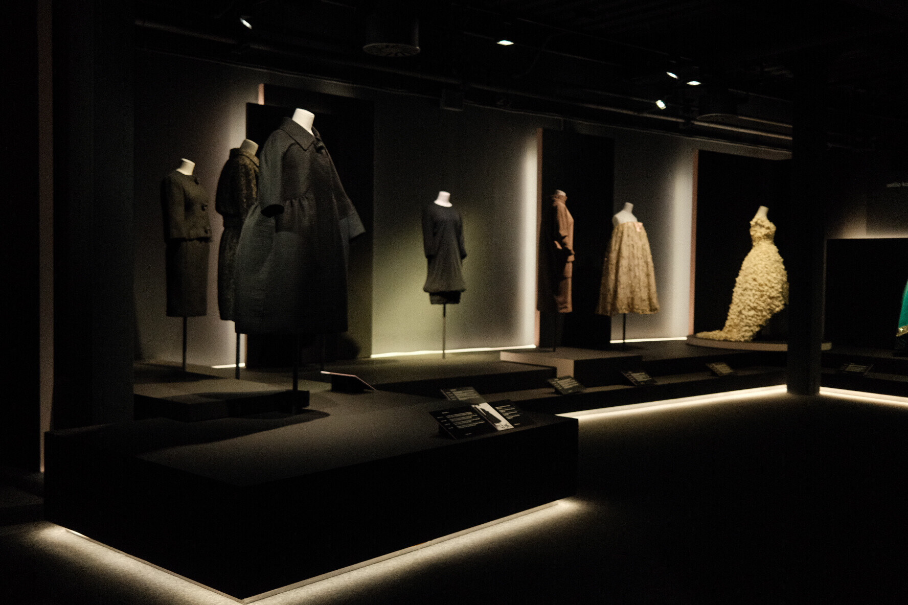 Dark museum gallery with seven dresses and coats on headless mannequins on black platforms under spotlights.