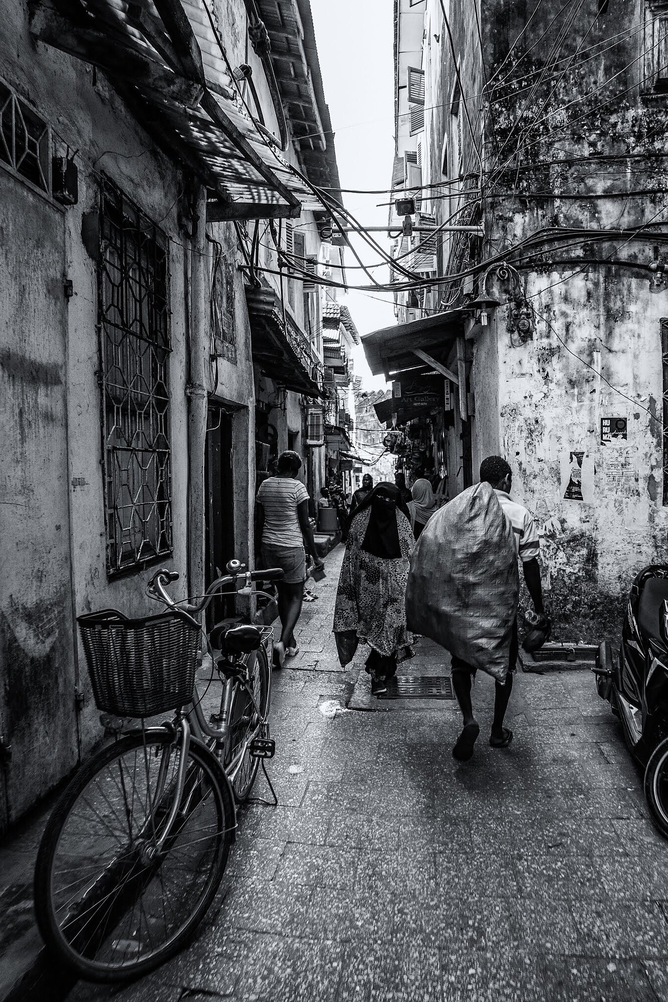Black-and-white photograph of a narrow wet alley, a bicycle with a basket leaning against the left wall, a man walking away carrying a large sack and a woman in a patterned dress ahead.